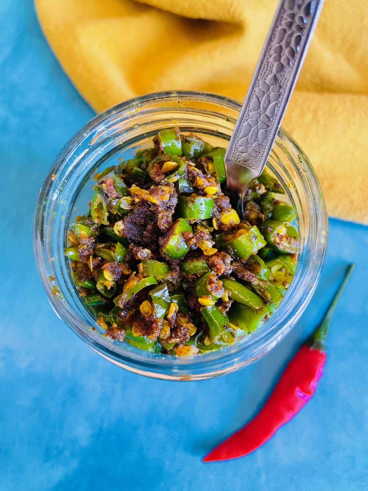 Top view of Mauritian Green Chili Achard in a glass jar and a stainless steel spoon inside the jar. A red Thai chili is in front of the jar.