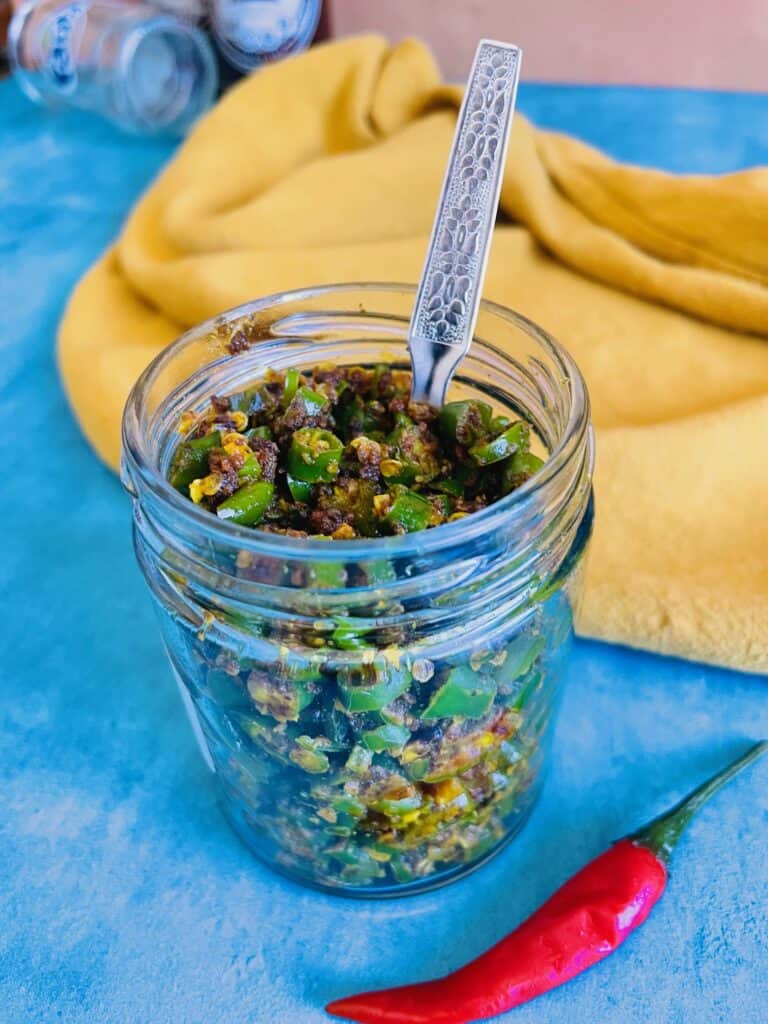 Mauritian Green Chili Achard in a glass jar and a stainless steel spoon inside the jar. A red Thai chili is in front of the jar.