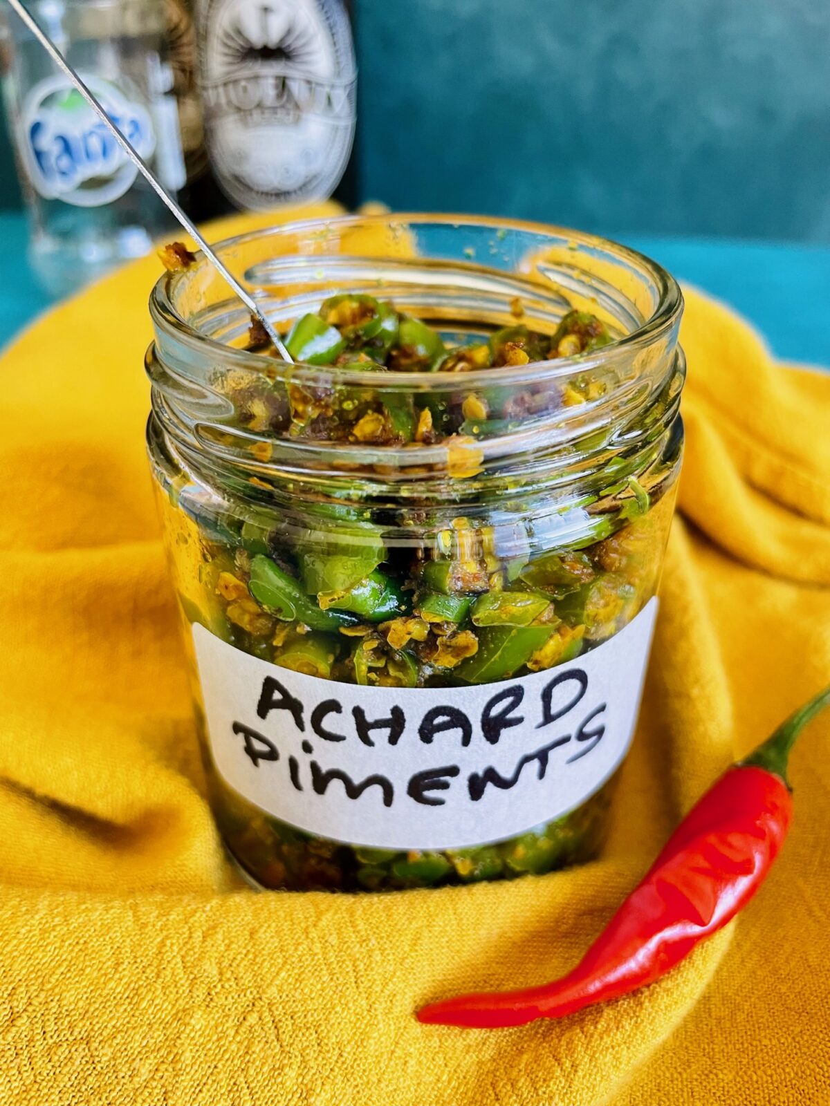 Mauritian Green Chili Achard in a glass jar and a stainless steel spoon inside the jar. A red Thai chili is in front of the jar.