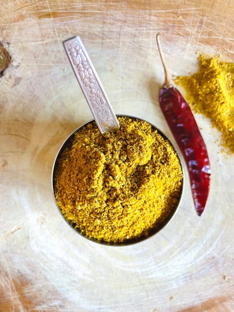 Mauritian Curry Powder in a stainless steel bowl and mini stainless steel spoon on chopping board.