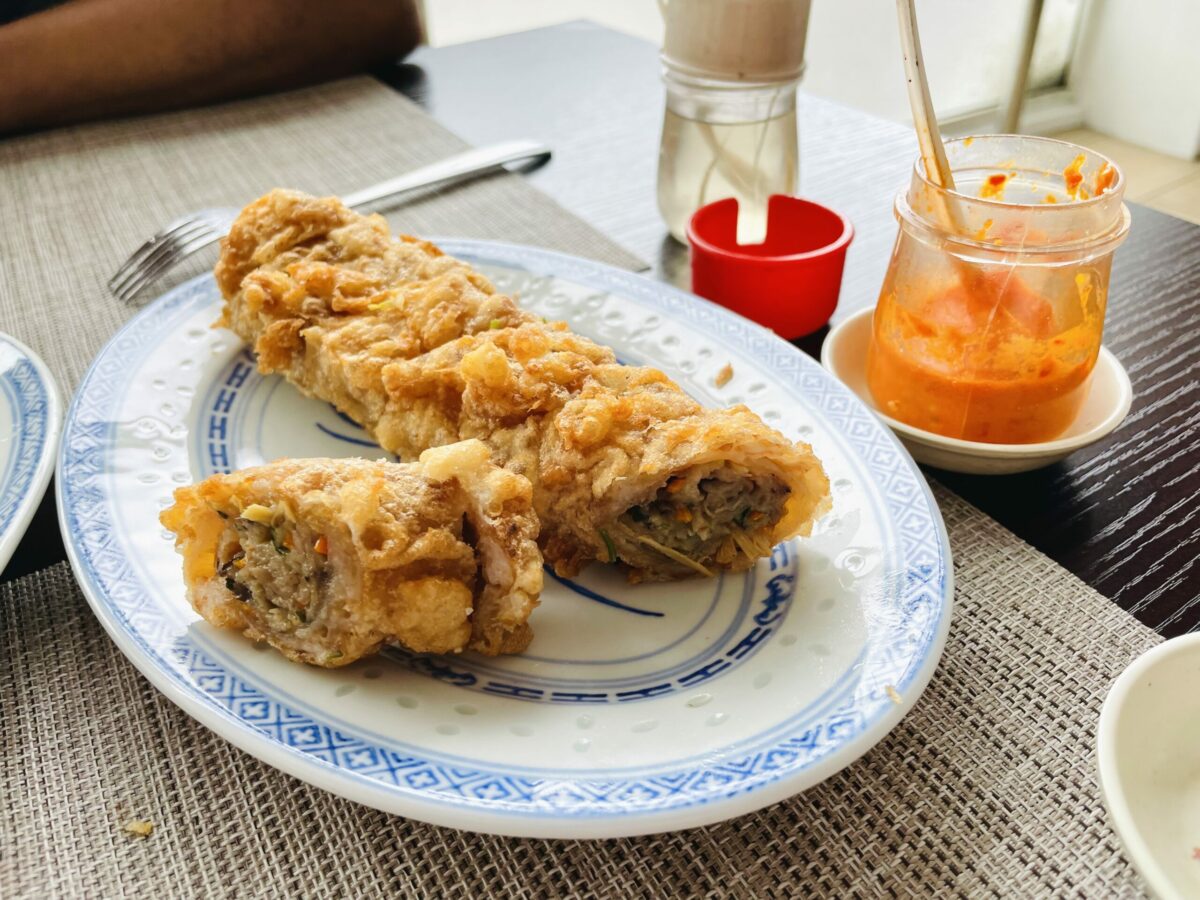 Chopped Mauritian hakien, rouleau de printemps, on Chinese blue dragon and white porcelain oval plate. Red chili sauce and garlic sauce condiments in bottles in the background.