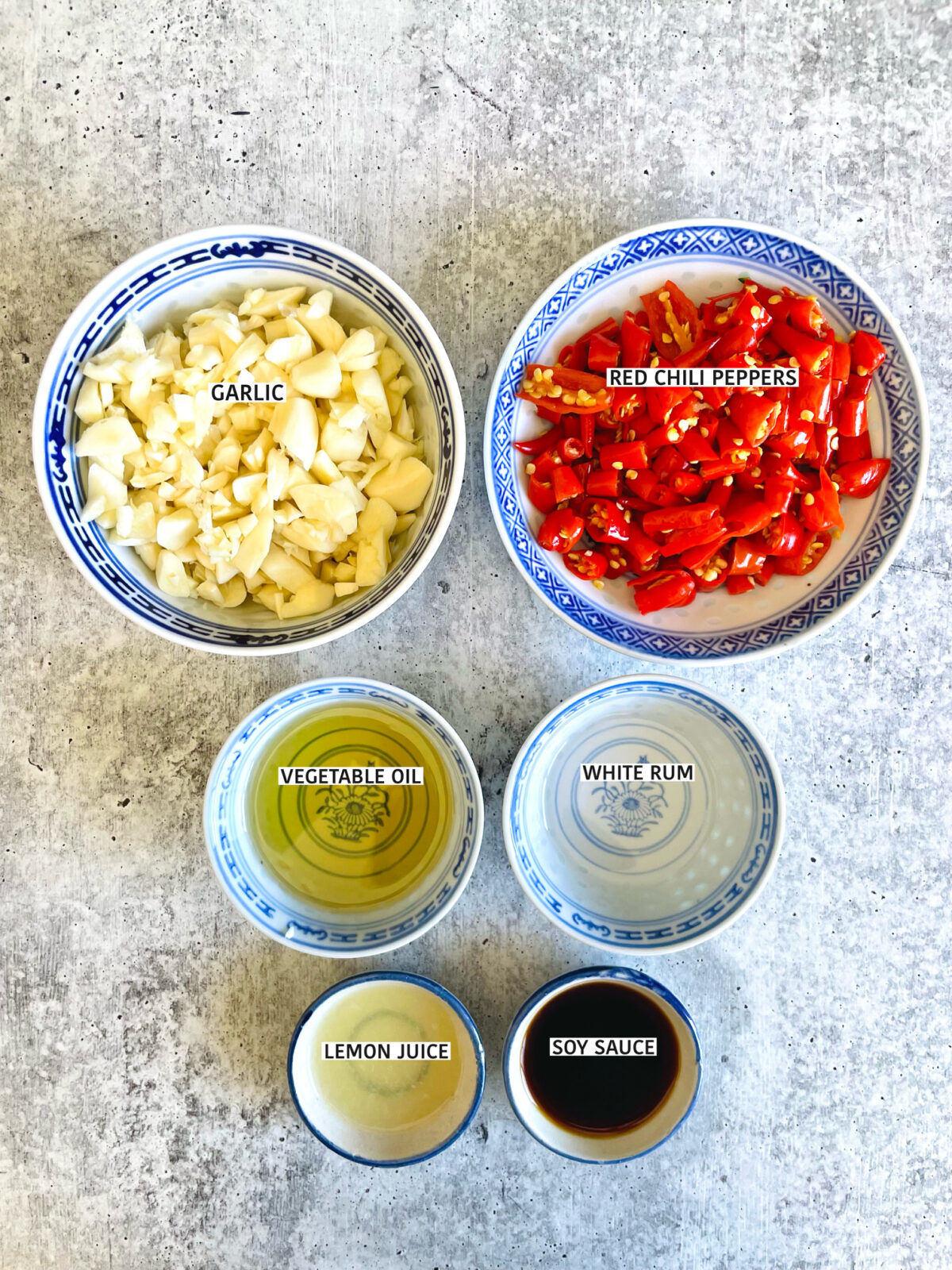 Chinese red chili paste mise en place in individual blue and white porcelain Chinese bowls with written labels: chopped garlic, chopped Thai chili peppers, vegetable oil, white rum, lemon juice and soy sauce.