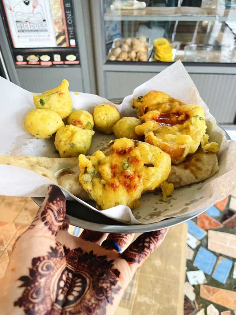 Henna-tattooed hand holding stainless steel plate of Mauritian gato de l'huile and folded roti: Bajia, bread pakora and eggplant pakora - Big Boss Vendor in Centre de Flacq