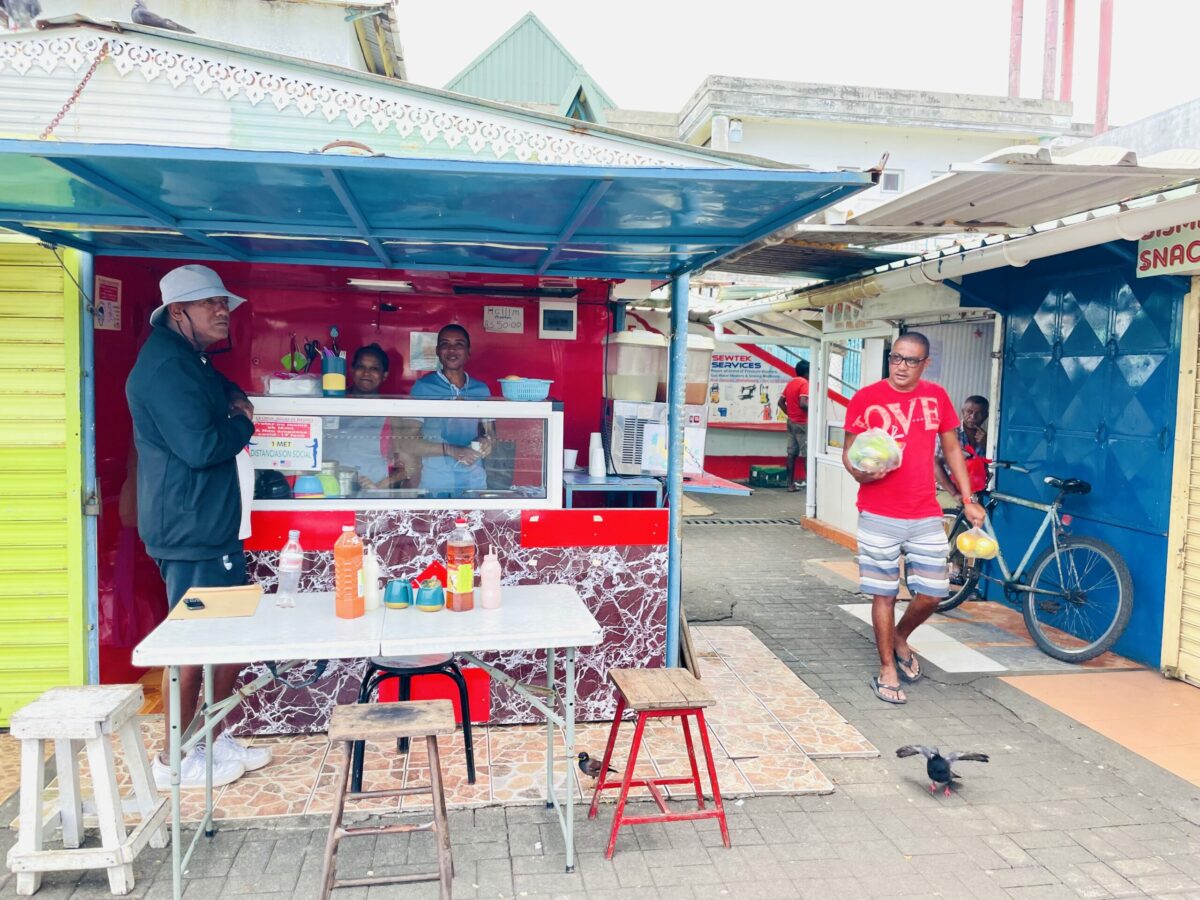 Halim Vendors in Mahébourg Market. Table and four different chairs in front of halim stall and a customer waiting.