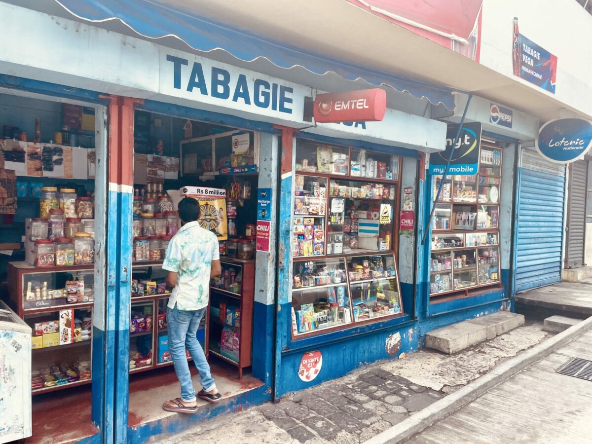 The front of Tabagie Mr. Vega with Mauritian snacks and candies and brick sidewalk. One customer standing inside the shop.