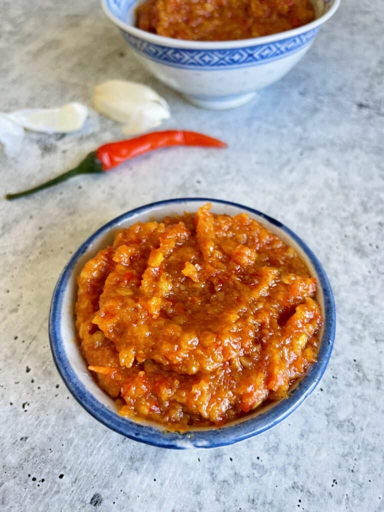 Mauritian Chinese red chili paste with fresh Thai red chili pepper, fresh garlic cloves and another small bowl with chili paste in the background.