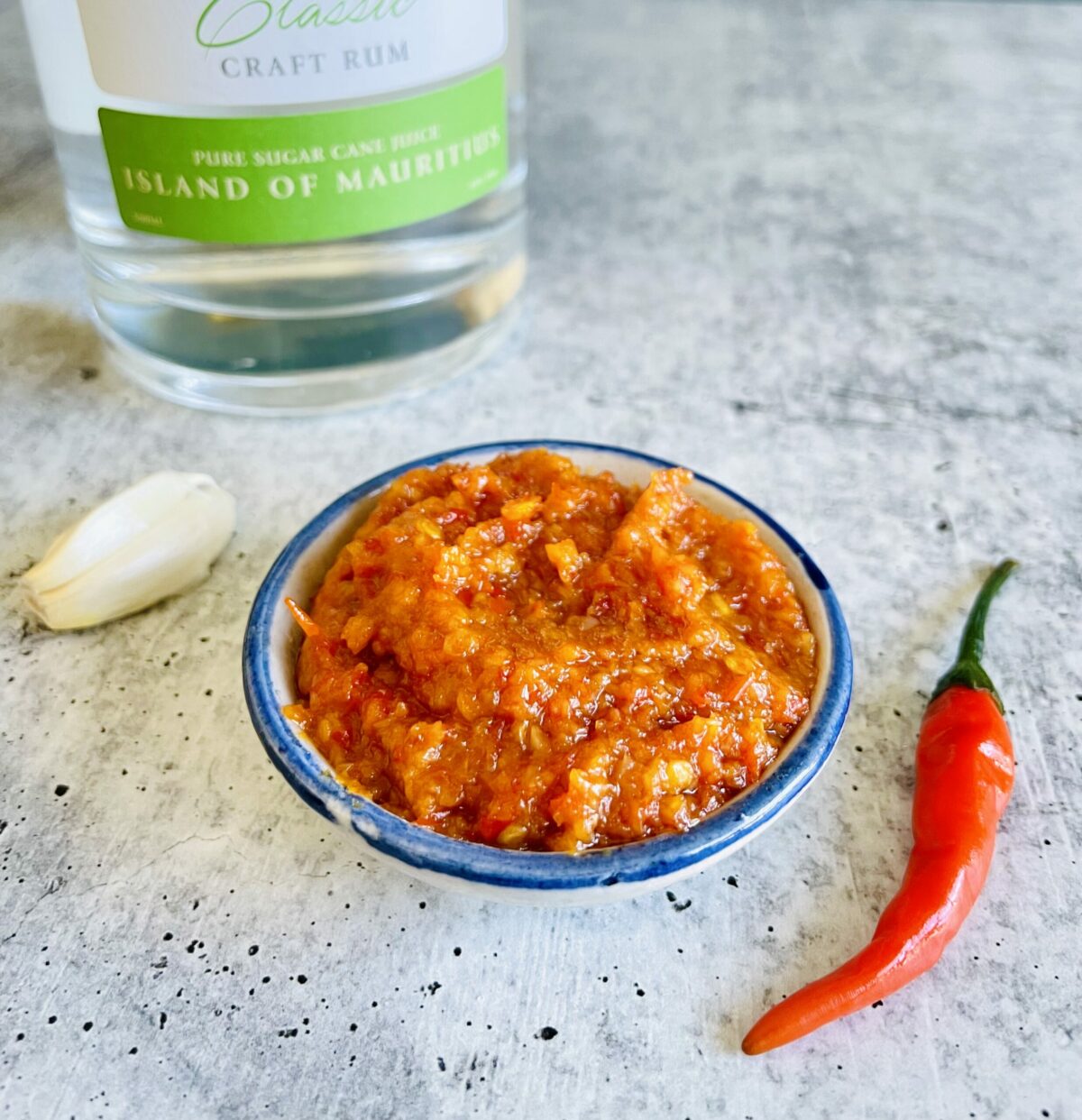 Mauritian Chinese red chili paste with white rum bottle in the background. Fresh Thai red chili pepper in the front right and two fresh garlic cloves on the left.