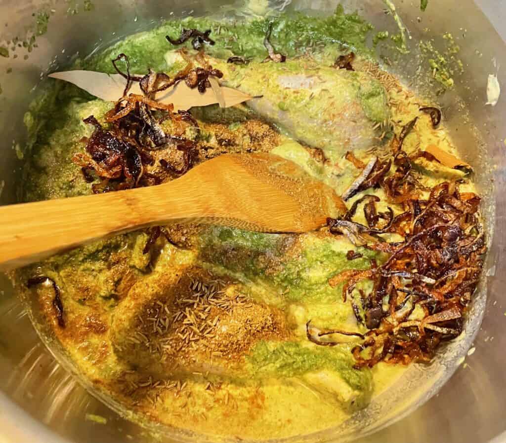 Marinaded chicken thighs and drumsticks mixed with briani sauce, peas and fried red onion in stainless steel stockpot. Wooden spatula stirring the mixture.