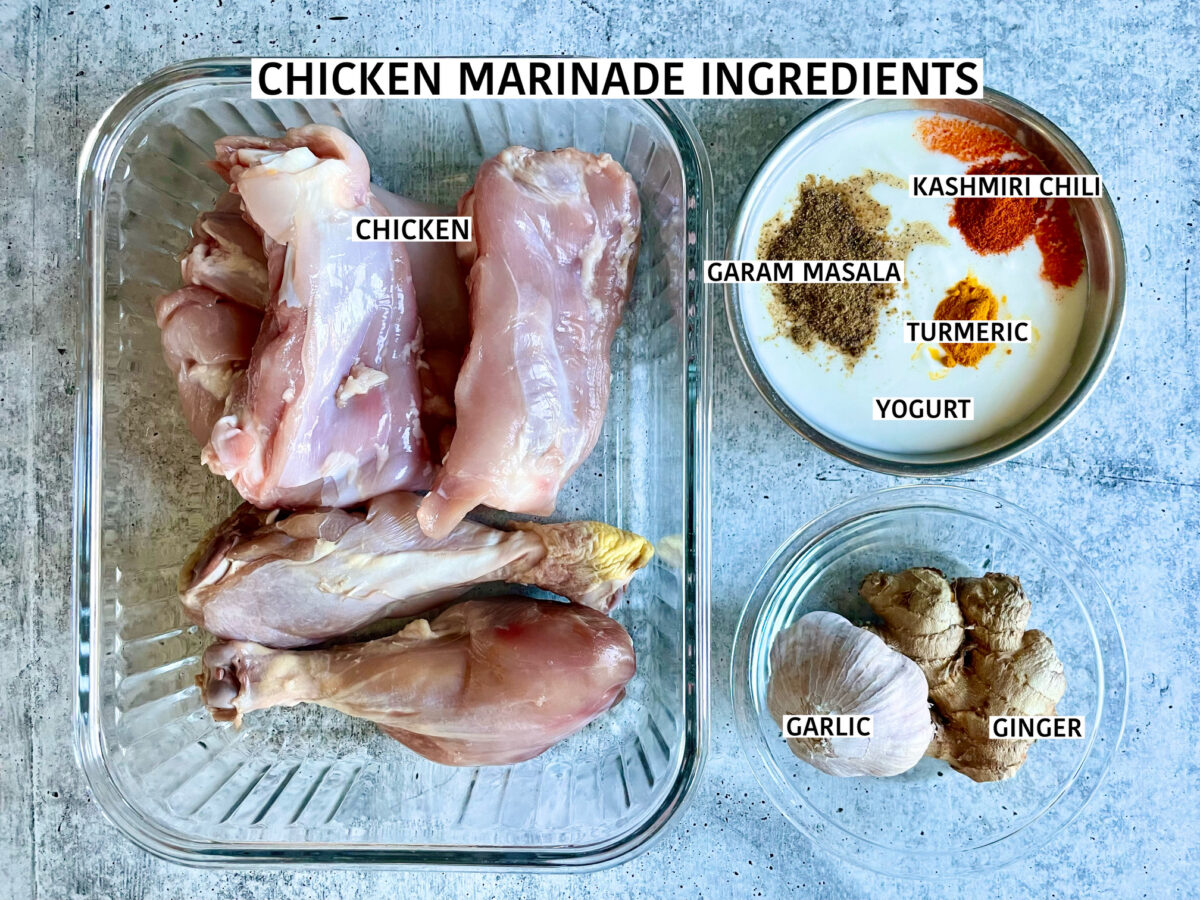 Ingredients for chicken marinade in individual bowls: chicken thighs and drumsticks, full-fat yogurt topped with garam masala, turmeric, Kashmiri chili, and a bowl of whole garlic bulb and ginger.