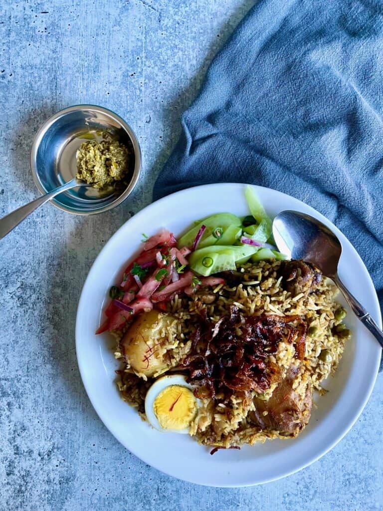 Plate of chicken briani with tomato chutney, cucumber salad, and topped with fried red onion. Spoon on the right of the plate. A small bowl of green chili paste with a teaspoon on the upper left side of the plate.