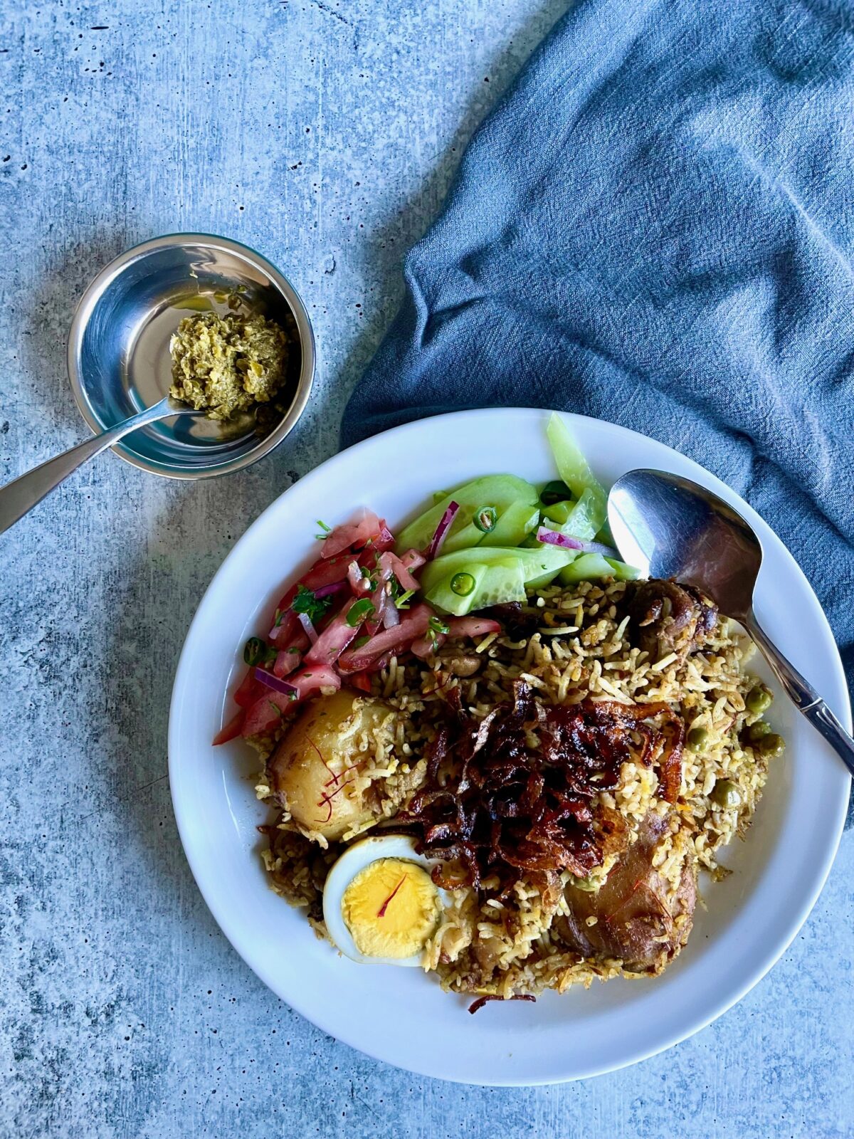 Plate of chicken biryani (briani) with tomato chutney, cucumber salad, and topped with fried red onion. Spoon on the right of the plate. A small bowl of green chili paste with a teaspoon on the upper left side of the plate.