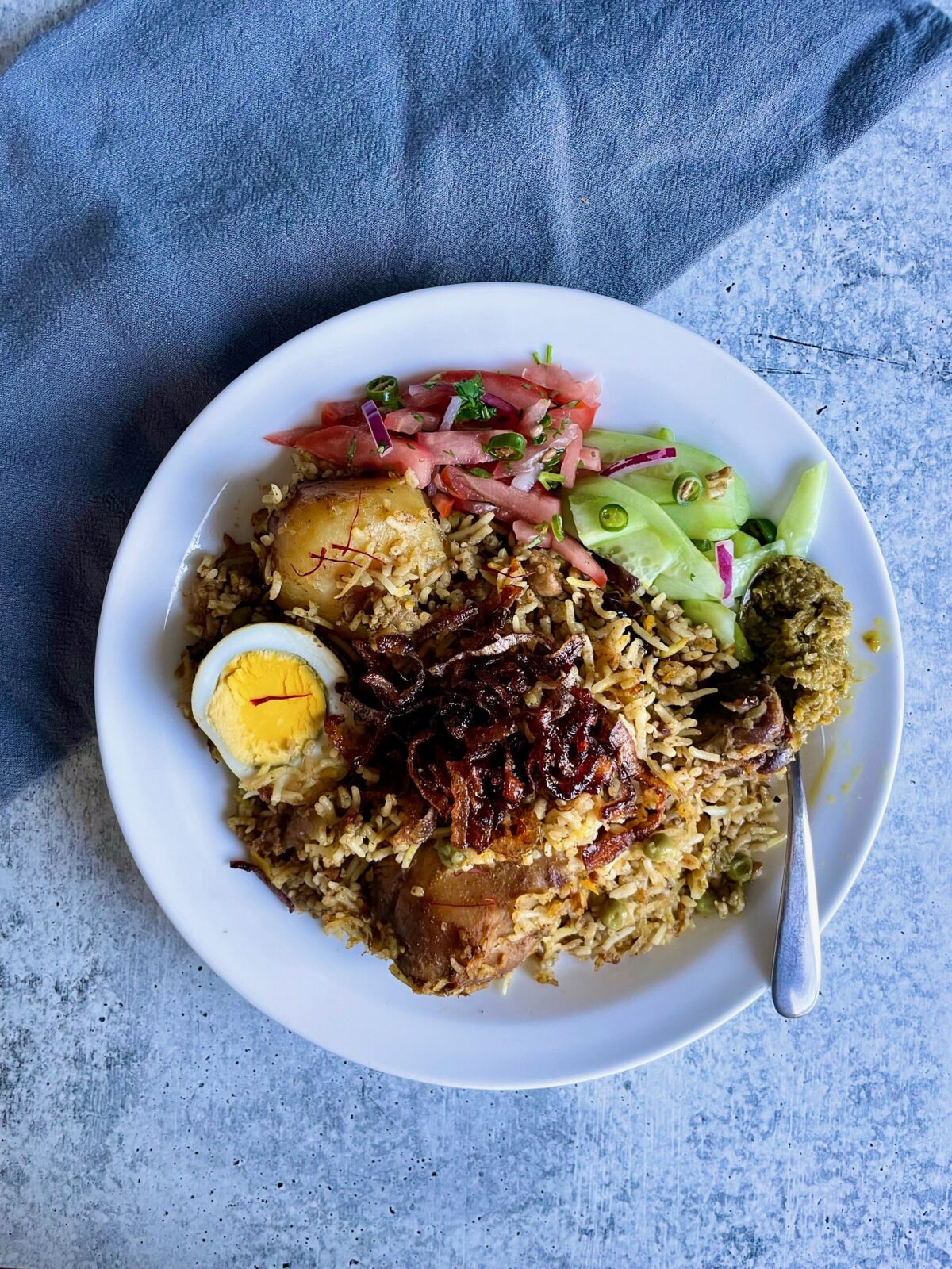 A plate of chicken briani with tomato chutney, cucumber salad, green chili paste, and topped with fried red onion. There is a blue towel on the upper left of the plate.