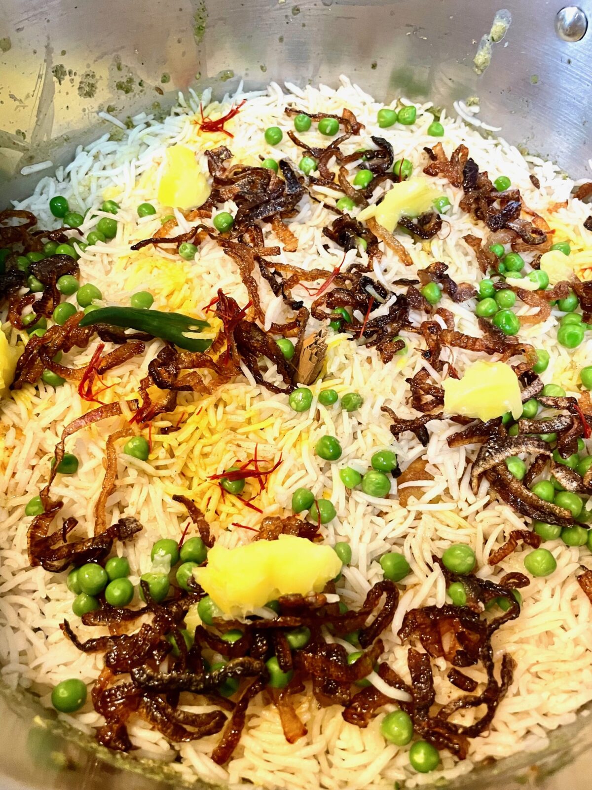 Top of chicken briani pre-cooking. Partially cooked basmati rice is topped with green peas, fried red onion, cultured ghee, green chili and saffron.