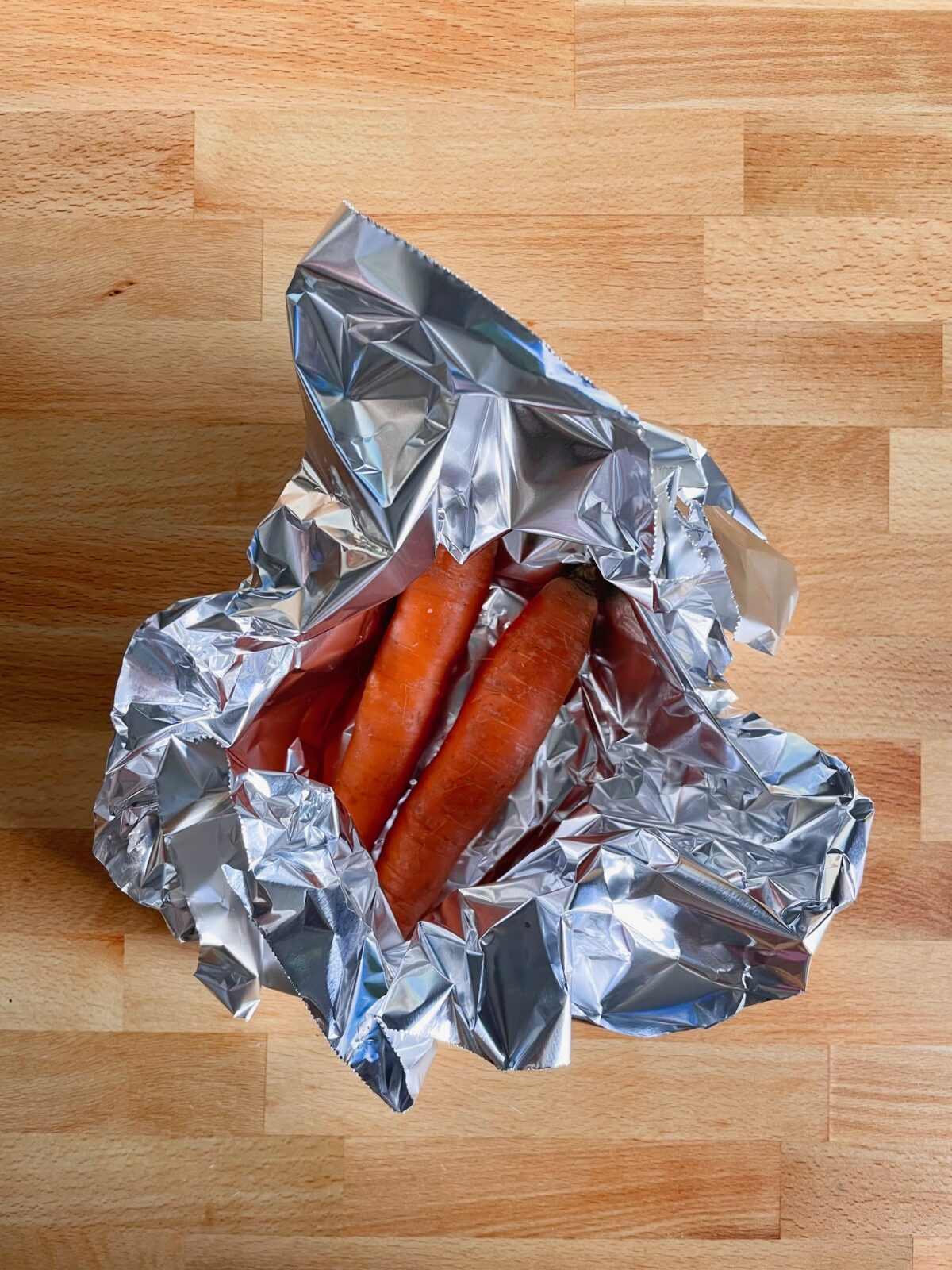 Two orange carrots partly wrapped in aluminum foil, and on a wooden board.