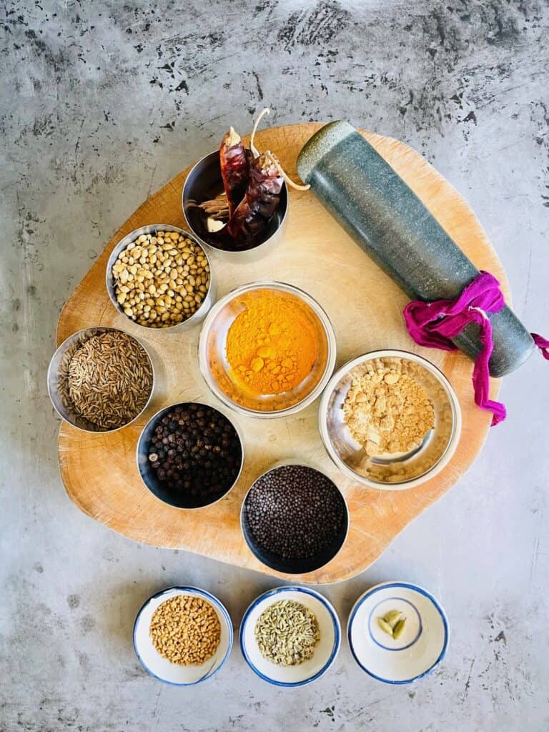 Whole spices for Indian curry powder in bowls: whole red chilies, cinnamon sticks, coriander seeds, cumin seeds, black peppercorn, turmeric powder, ginger powder, mustard seeds, fenugreek seeds, fennel seeds and cardamom pods.
