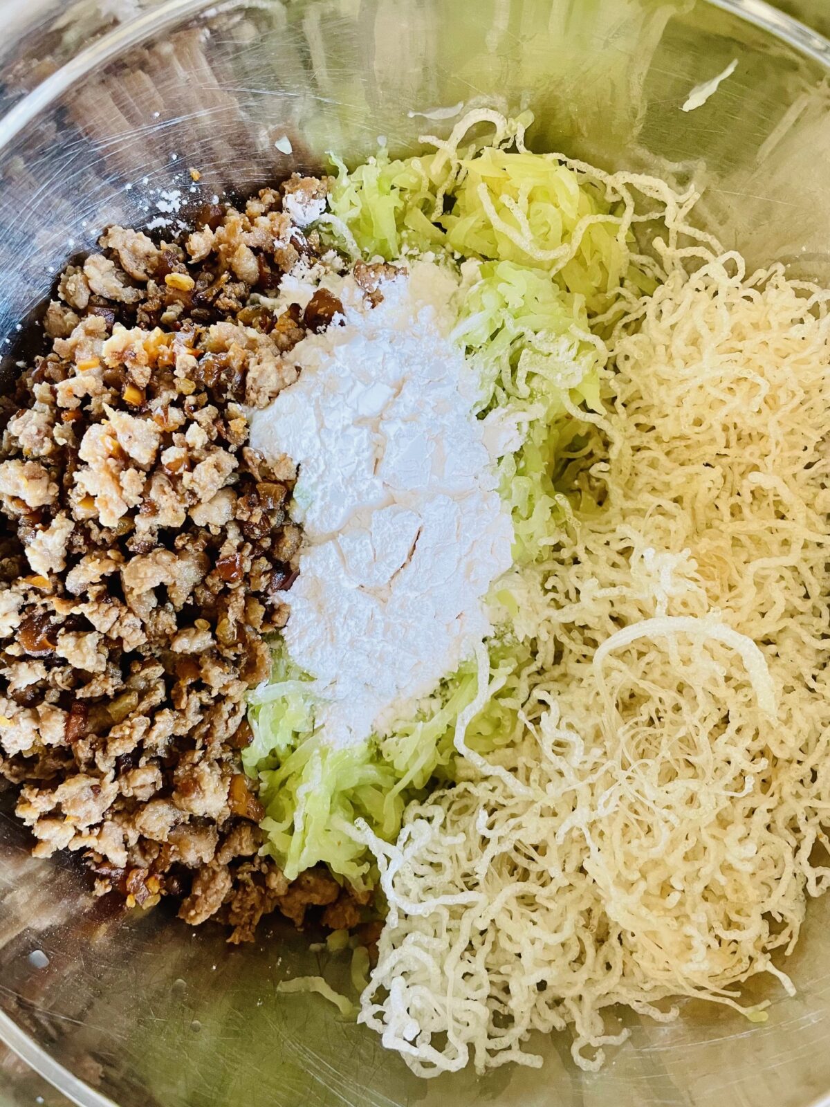 Boulette chouchou ingredients in stainless steel bowl. From left to right: chicken mixture, grated chayote, fried rice vermicelli. All topped with tapioca flour.