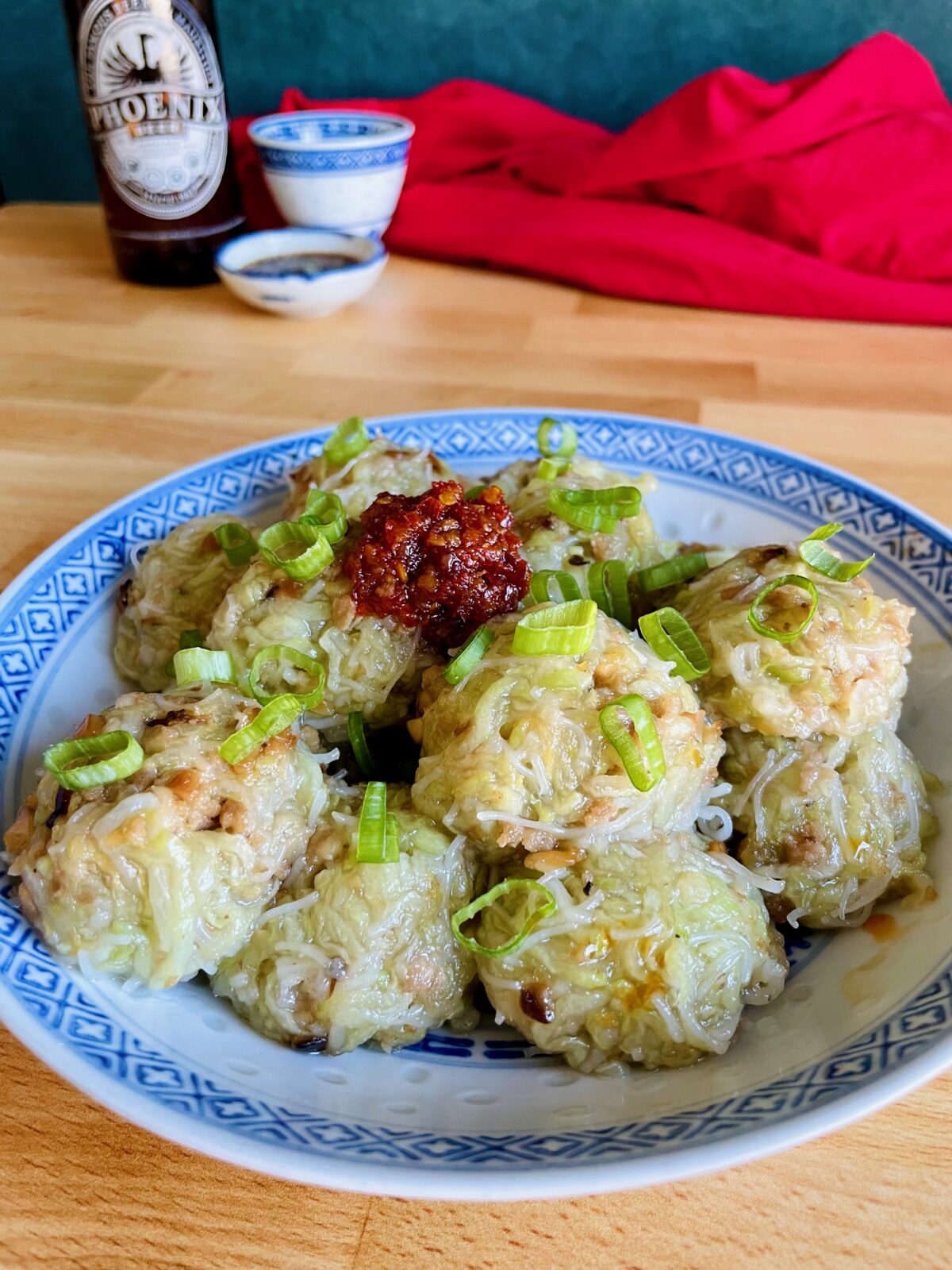 Boulette chouchou in plate topped with red chili paste and green onion. Bottle of Mauritian Phoenix beer in the background and mini Chinese bowls. Red cloth next to the bowls.