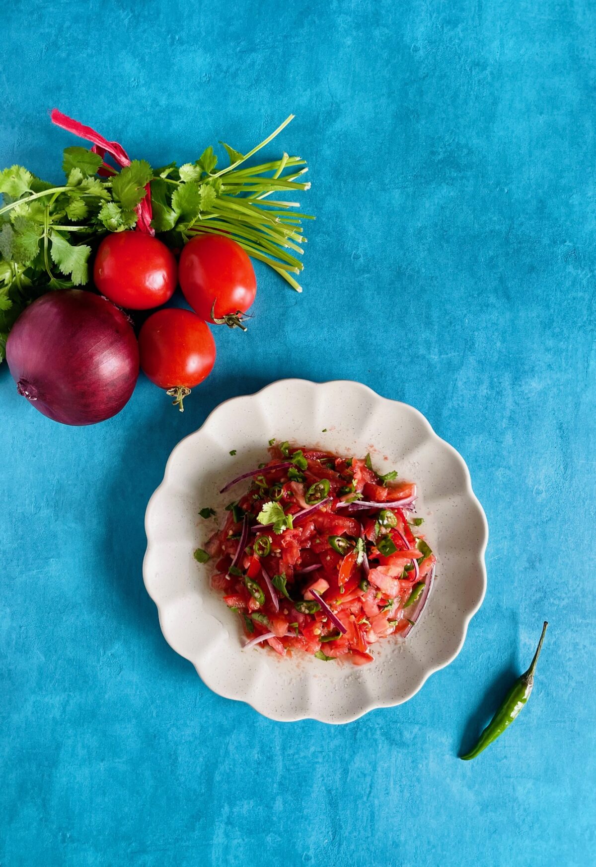 Satini pomme d'amour (Mauritian tomato salad). Sliced tomatoes, chopped green chilies, sliced red onion in a plate with scalloped edges. Whole red onion, bunch of cilantro, and tomatoes in the background.