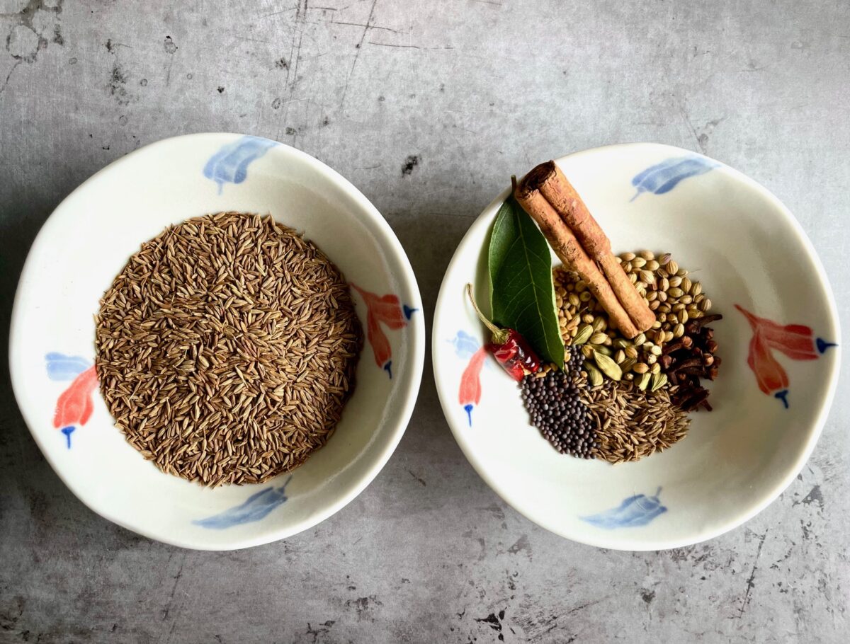 Cumin seeds and whole spices for curry powder in bowls. Whole spices: curry leave, red chili, cinnamon, brown mustard seeds, cloves, cardamom pods, fenugreek seeds, cumin seeds, and coriander seeds.