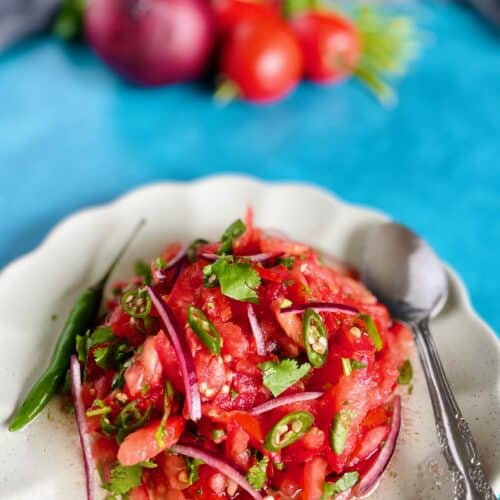 Mauritian tomato chutney with a whole green chili, sliced tomatoes, chopped green chilies, sliced red onion in a plate with scalloped edges. Spoon on the edge of the plate.