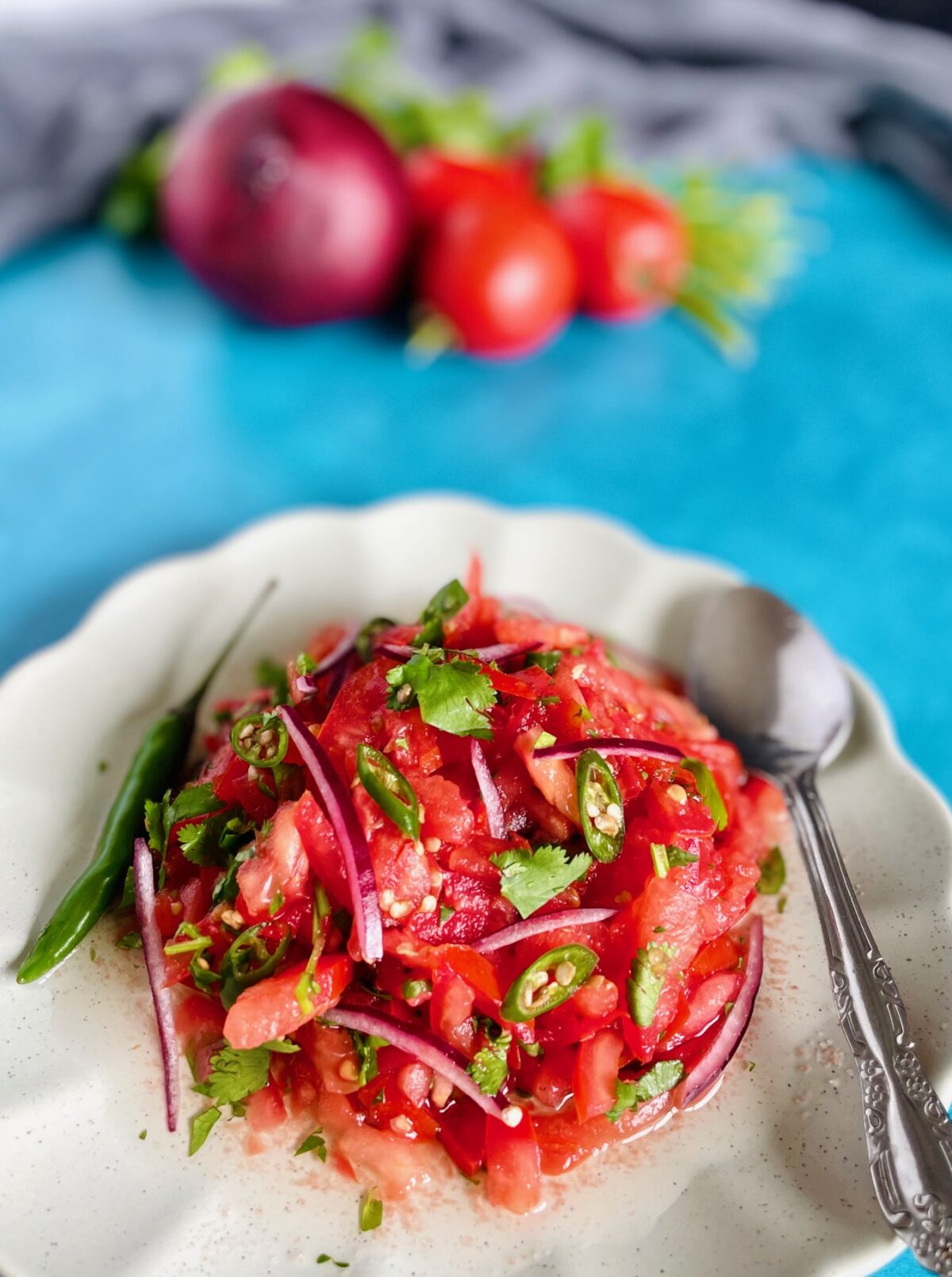Mauritian tomato chutney with a whole green chili, sliced tomatoes, chopped green chilies, sliced red onion in a plate with scalloped edges. Spoon on the edge of the plate.