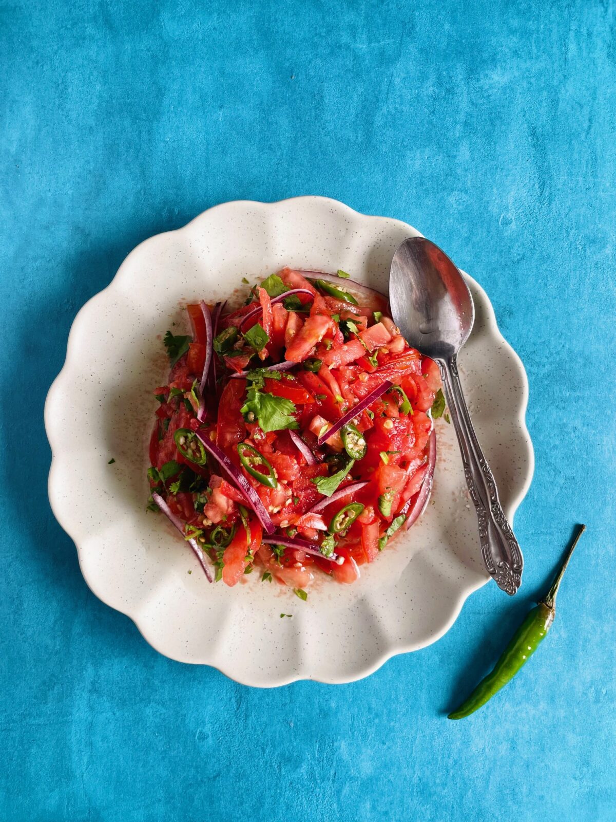 Satini pomme d'amour avec piment. Sliced tomatoes, chopped green chilies, sliced red onion in a plate with scalloped edges. Spoon on the edge of the plate.